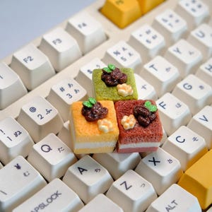 May include: Three miniature, colorful cake-shaped keycaps rest on a white keyboard. The keycaps are square, with layers of orange, yellow, and red, topped with brown and green decorative elements. The keyboard has white keys with Japanese characters.