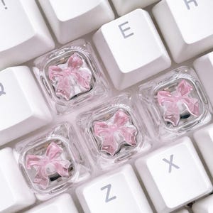 May include: Three square, transparent mechanical keyboard keycaps, each featuring a small, sculpted, light pink bow embedded within the clear resin. These decorative keycaps are positioned among standard white keys on a computer keyboard.