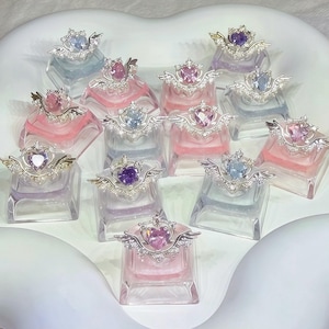 Pode incluir: Uma coleção de pequenos frascos de perfume decorativos. Cada frasco tem uma base retangular transparente com um líquido colorido no interior. As tampas apresentam um design prateado em forma de coração com asas e uma coroa, adornado com pequenos cristais transparentes. Os frascos estão dispostos numa superfície branca.