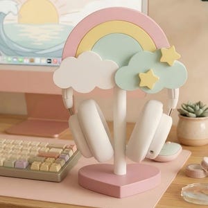 May include: A pastel-colored headphone stand shaped like a rainbow with clouds and stars. The stand is pink at the base and has a white central support. White headphones are hanging on the stand. A keyboard and computer are in the background.
