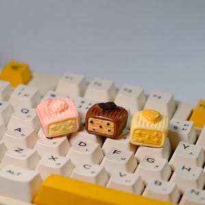 May include: Three decorative keyboard keycaps shaped like miniature cakes. One is pink with a white topping, another is brown with a chocolate accent, and the third is yellow with a cheese-like topping. The keycaps sit on a white keyboard.