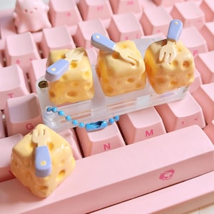 Peut inclure: Ensemble de quatre touches de clavier jaunes en forme de fromage, ornées de fourchettes bleues. Elles sont fixées à une base en plastique transparent avec une chaîne de perles bleues, sur un clavier rose.