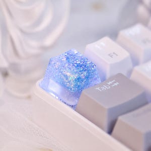 Puede incluir: Una tecla mecánica cuadrada con un acabado de resina azul translúcida incrustada con partículas de purpurina iridiscente. La tecla está colocada sobre una base de teclado blanca junto a una tecla gris etiquetada con la palabra Tab.