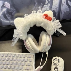 May include: White headphones with pink accents are paired with a fluffy white cat ear headband adorned with red pom-poms and lace. A keyboard and mouse with manga-style art are also visible.