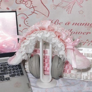 May include: Grey over-ear headphones adorned with a decorative, ruffled headband cover featuring layers of white lace and soft pink fabric, accented with small pink satin bows on each side, resting on a white U-shaped headphone display stand.