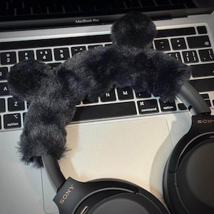 May include: Black fluffy microphone cover and headphones resting on a laptop keyboard. The headphones are black with the brand name "SONY" visible on the earcups. The laptop is a MacBook Pro.