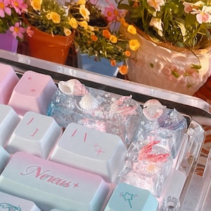 May include: A mechanical keyboard featuring custom resin keycaps with embedded seashells, pearls, and mermaid tail motifs. The keys include white and pastel gradient caps with the text Nereus and star symbols, set against a clear, transparent frame.
