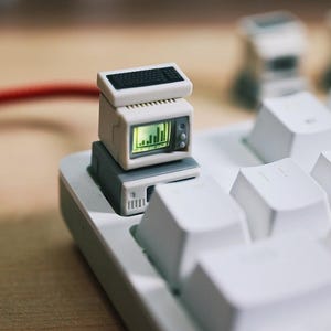 May include: A miniature computer keycap featuring a small, retro-style computer monitor with a keyboard on top. The monitor displays a green bar graph. The keycap is white and grey, and sits on a white keyboard.