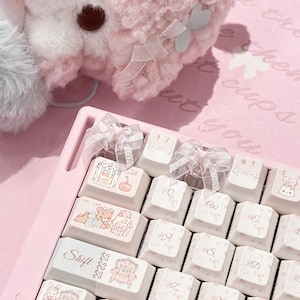 May include: A light pink mechanical keyboard featuring white keycaps with various illustrations including a teddy bear, a rabbit, and furniture. Two keycaps are adorned with sheer white ribbon bows. The shift key displays the text Shift.