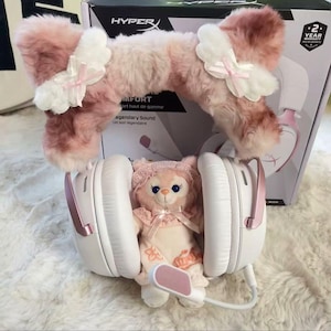 Può includere: Cuffie da gioco bianche e rosa con un accessorio per l'orecchio in peluche rosa. Le cuffie sono dotate di microfono e sono abbinate a un peluche con un cappello rosa. La scatola dietro le cuffie riporta la scritta "HYPER".