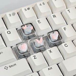 Puede incluir: Primer plano de un teclado blanco con teclas personalizadas. Las teclas transparentes presentan diseños de mariposas y corazones rosas y azules. Las teclas del teclado incluyen Q, E, R, T, F, C, Z, X y Alt.