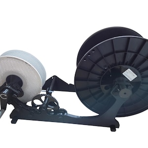 May include: A black and silver 3D printer filament spool holder with a hand crank and gears. The holder has two large spools, one silver and one black, mounted on a black base. The gears and crank suggest manual operation.