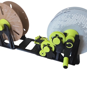 May include: A black and lime green 3D printer filament spool winder featuring a manual hand crank, a series of interlocking gears, and two circular spool holders. One spool is light brown, and the other is white with a perforated surface pattern.
