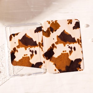 Może przedstawiać: Przezroczyste etui na telefon z nadrukiem krowy. Etui jest otwarte, odsłaniając wnętrze i pasujący uchwyt na karty z nadrukiem krowy. Nadruk krowy ma brązowe i beżowe plamy na kremowym tle.