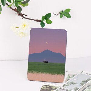 May include: A rectangular art print featuring a landscape scene. The image depicts a small cabin on a green field, a mountain, and a sunset sky with a moon. The print has a rounded edge and a light brown border.