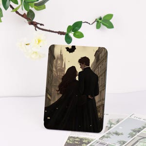 May include: A rectangular case featuring a romantic illustration of a couple in black attire, set against a European cityscape with a bat silhouette. The artwork has a sepia tone, evoking a vintage aesthetic.