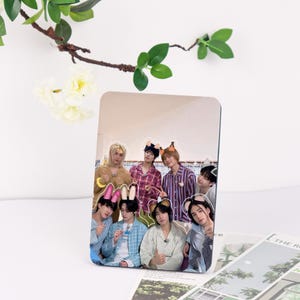 May include: A rectangular photo print showing seven people in pyjamas and animal ear headbands. The image has a glossy finish and is set against a white background with decorative greenery. The print is likely a fan item.