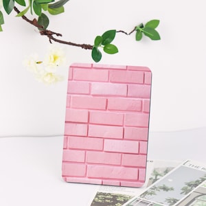 May include: A pink rectangular object with a brick pattern stands upright. The object is a light pink colour, with darker pink lines defining the brick shapes. Green leaves and white flowers are in the background.
