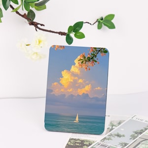 May include: Rectangular decorative item featuring a serene seascape with a sailboat under a cloudy sky. The image has a vibrant color palette of blues, oranges, and whites. A branch with green leaves and small white flowers is in the upper left corner.