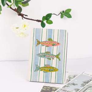 May include: A notebook with a striped blue and white background featuring three colorful fish illustrations. The fish are pink, blue-green, and yellow with detailed fins and scales. The notebook is a decorative item.