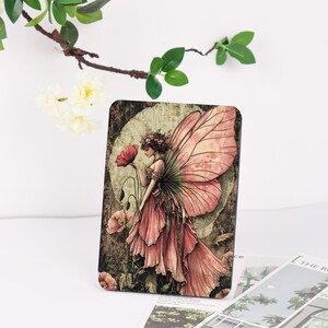 May include: A rectangular art print featuring a fairy with pink butterfly wings and a flowing pink dress, holding a red poppy. The background has a vintage, antique aesthetic with a circular design and a dark, textured border.