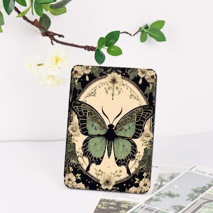 May include: A rectangular decorative item featuring a detailed illustration of a butterfly with green and black wings. The butterfly is set against a cream-colored circle, surrounded by floral and botanical elements on a black background.