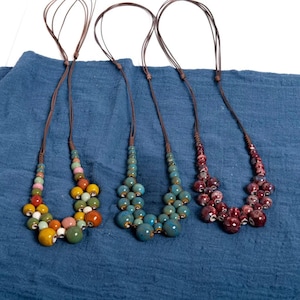 May include: Four handmade necklaces with ceramic beads in various colors. The necklaces have brown leather cords and feature clusters of colorful beads in shades of yellow, green, pink, orange, blue, and burgundy. The necklaces are displayed on a blue fabric background.