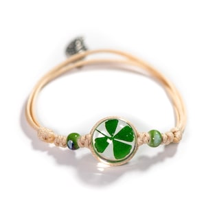 Puede incluir: Un brazalete de cuerda beige con un dije de trébol de cuatro hojas verde en un círculo de vidrio transparente. El brazalete tiene dos cuentas de vidrio verde a cada lado del dije.