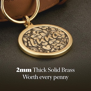 May include: A round solid brass pet tag featuring an engraved floral pattern and the name LUNA in the center. The tag is attached to a gold-toned metal ring and is 2mm thick. The text 2mm Thick Solid Brass Worth every penny is visible.