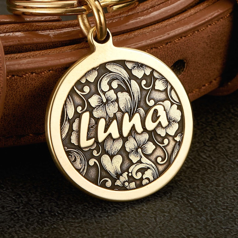 May include: A round gold-toned metal pet ID tag featuring a raised floral and scrollwork pattern with a dark recessed background. The name Luna is engraved in large, smooth, light-colored script letters across the center of the circular tag.