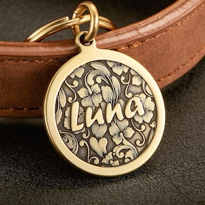 May include: A round gold-toned metal pet tag featuring an embossed floral pattern and the name Luna in raised script lettering. The tag is attached to a gold-toned split ring, which is looped through a brown leather collar with visible stitching.