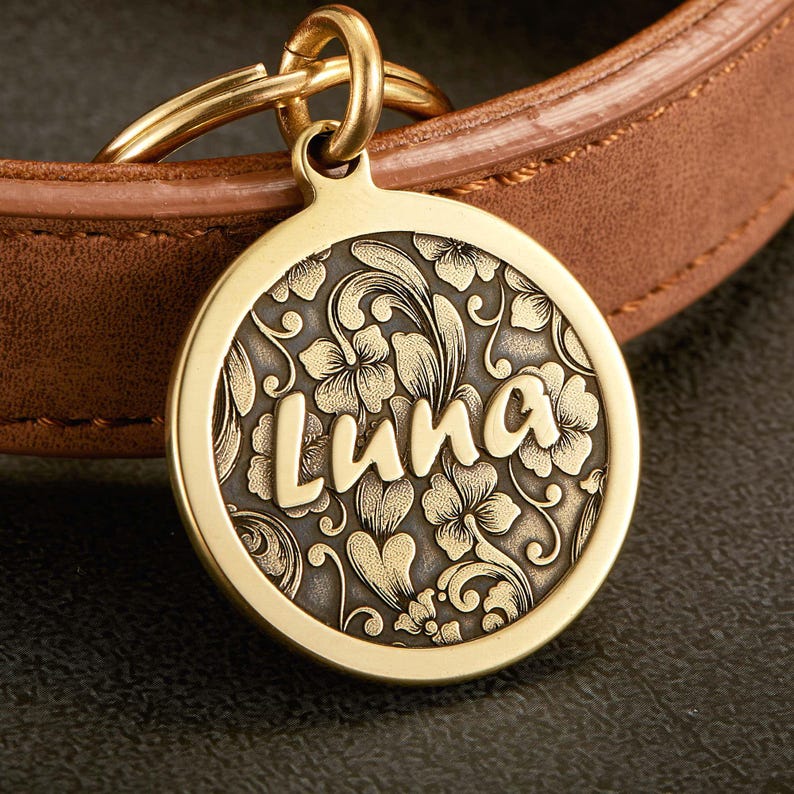 May include: A round gold-toned metal pet tag featuring an embossed floral pattern and the name Luna in raised script lettering. The tag is attached to a gold-toned split ring, which is looped through a brown leather collar with visible stitching.