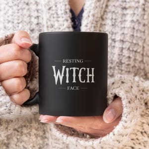 May include: A black mug with the words "RESTING WITCH FACE" in white lettering. The mug is held by a person wearing a light-coloured sweater. The mug has a black handle.