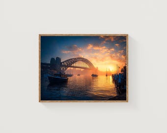 May include: A framed photograph of the Sydney Harbour Bridge at sunrise. The bridge is silhouetted against a vibrant orange and yellow sky. Boats are in the water, and the image has a warm, inviting feel.