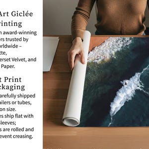 a person carefully handling a roll of paper, possibly for art print packaging. The person is standing behind a wooden table, which has a large piece of paper on it. The paper appears to be a watercolor print of a seascape, with waves crashing against the shore. The person is holding the paper up to the light, possibly to examine it or prepare it for printing.