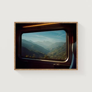 Può includere: Una fotografia incorniciata cattura una vista panoramica delle montagne da un finestrino del treno. Il paesaggio presenta strati di colline e montagne verdi sotto un cielo blu. La cornice della finestra è scura e l'interno del treno è visibile.