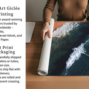 a person carefully handling a roll of paper, possibly for art print packaging. The person is standing behind a wooden table, which has a large piece of paper on it. The paper appears to be a watercolor print of a seascape, with waves crashing against the shore. The person is holding the paper up to the light, possibly to examine it or prepare it for printing.