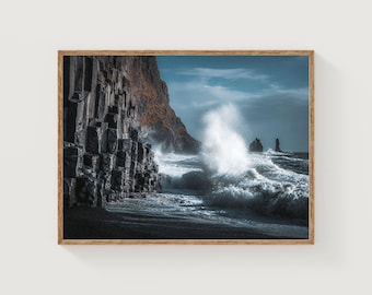 May include: A framed print depicting a dramatic coastal scene. A large wave crashes against a dark, rocky shore with distinctive columnar rock formations. The sky is a blend of blue and grey.