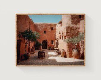 Peut inclure: Une impression encadrée d'une cour avec des bâtiments couleur terre cuite et un ciel bleu clair. La cour comprend des plantes en pot, de petites tables et de grands pots en argile. L'image évoque une sensation de chaleur et de tranquillité.