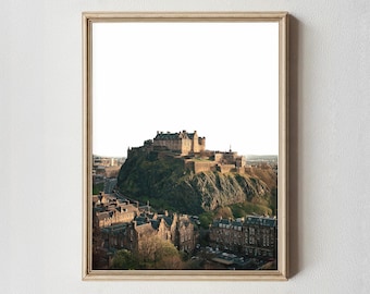 May include: A framed print featuring Edinburgh Castle atop a hill, surrounded by buildings. The castle is a large, stone structure with multiple towers and battlements. The print has a light-colored wooden frame.