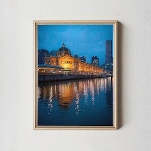 May include: A framed print showing a building at dusk, with a domed roof and lit windows. The building's lights reflect in the water, with a city skyline in the background. The sky is a deep blue.