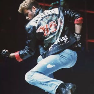 May include: A performer in a black leather jacket with red trim, light blue jeans, and black boots. The jacket has a decorative back with the text "Screws" and "85" in red. The performer is in a dynamic pose.