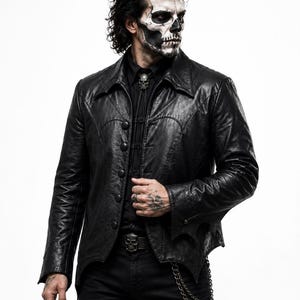 May include: A black leather jacket with a pointed hem, long sleeves, and a button-up front. The jacket features a structured collar and curved seam detailing across the chest. It is worn over a black button-down shirt with a silver skull bolo tie.