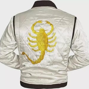 May include: A white quilted satin bomber jacket featuring a large, detailed gold scorpion embroidery on the back. The jacket has a dark brown ribbed collar, cuffs, and hem, with a diamond-stitched pattern across the white fabric body.