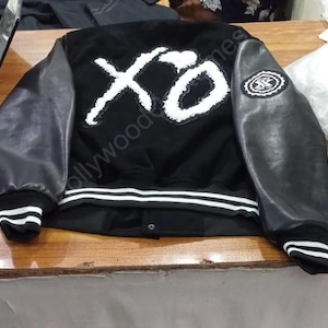 May include: A black varsity jacket featuring long black sleeves, a large white fuzzy XO logo on the back, and a smaller circular white patch on the right sleeve. The jacket includes white striped ribbed cuffs and a white striped hem at the bottom.