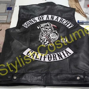 May include: A black leather vest featuring a large white embroidered patch on the back with the text Sons of Anarchy, a graphic of a grim reaper holding a scythe, a small MC patch, and a bottom rocker patch with the text California.