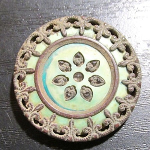 May include: A round, antique button with a light green center featuring a floral design. The center is surrounded by a bronze-colored ring and an ornate, openwork border. The button has a vintage aesthetic.