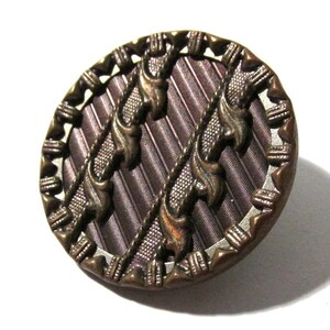 May include: A round, antique button with a dark brown and bronze color scheme. The button features a textured, ribbed center with decorative bronze accents and a detailed, patterned edge. The button is set against a white background.