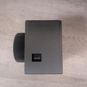 May include: A dark gray rectangular device with a USB port and a cylindrical, ridged component on the left side. The device is set against a light wood-grain surface.