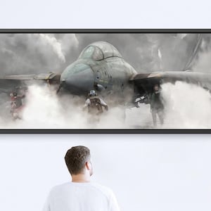 May include: A framed artwork depicting a military jet emerging from a cloud of smoke. The artwork is displayed on a white wall, with a person sitting on a blue bench in front of it.